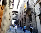 Menschen schlendern durch eine enge Gasse in der **Barcelona Altstadt**. Über ihnen spannt sich eine historische Brücke mit filigranen Bögen.