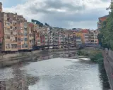Bunte Häuserfronten säumen den Fluss Onyar in der malerischen **Girona Altstadt**. Eine Brücke verbindet die Ufer, während sich die Wolken am Himmel spiegeln.