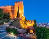 Die Alcazaba von Almería erstrahlt im Abendlicht. Beleuchtete Wege führen durch Gärten zur imposanten Festung.