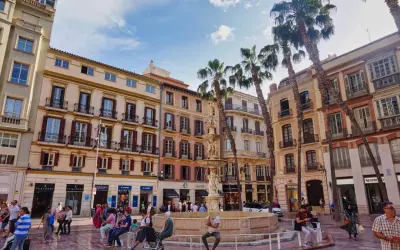 Sonniger Platz in der **Malaga Altstadt** mit einem Brunnen, Palmen und bunten Gebäuden. Menschen flanieren und genießen die Atmosphäre.