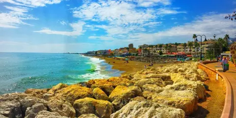 Sonniger Tag an der playa de la carihuela. Türkises Meer, goldener Sandstrand mit Liegen und Palmen, gesäumt von bunten Gebäuden. Felsen im Vordergrund.