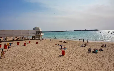 Sonniger Cadiz Strand mit Menschen, die sich am Meer entspannen. Ein weißes Gebäude mit Kuppel steht am Ufer.