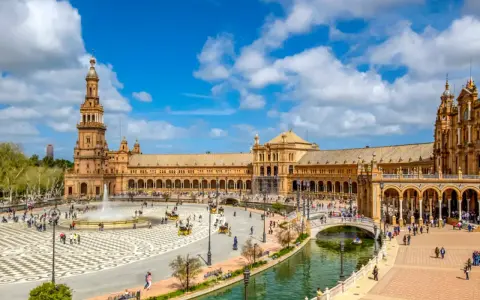 Der beeindruckende Sevilla Palast mit seinen Türmen und Arkaden, umgeben von einem Kanal und belebtem Platz mit Brunnen und Kutschen.