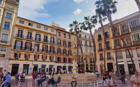 Sonniger Platz in der **Malaga Altstadt** mit einem Brunnen, Palmen und bunten Gebäuden. Menschen flanieren und genießen die Atmosphäre.