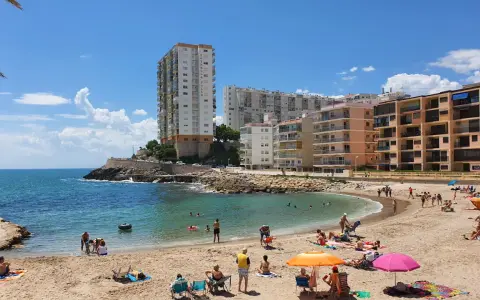 Sonniger Cullera Strand mit vielen Menschen, die sich im Meer und am Sand entspannen, im Hintergrund hohe Gebäude.