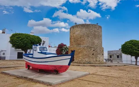 Ein Fischerboot und ein alter Turm in der conil altstadt. Weiße Häuser und grüne Bäume unter blauem Himmel mit Wolken.