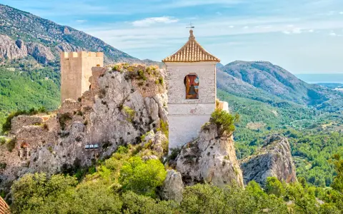 Das Castell de Guadalest thront auf einem Felsen, umgeben von grünen Bergen und dem blauen Meer in der Ferne. Ein Glockenturm ragt empor.