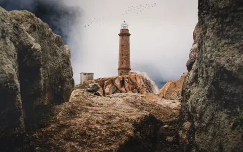 Leuchtturm auf felsiger Küste der Costa da Morte, Vögel ziehen über neblige Berge.