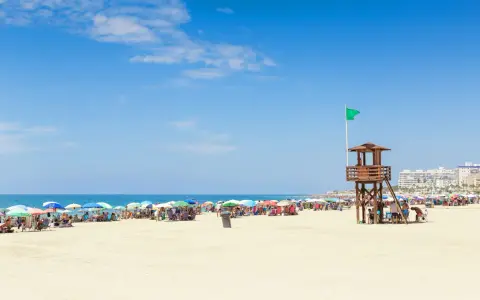 Sonniger Strand in Rota, Spanien, mit vielen bunten Sonnenschirmen und Menschen. Ein Rettungsschwimmerturm mit grüner Flagge steht am Meer.