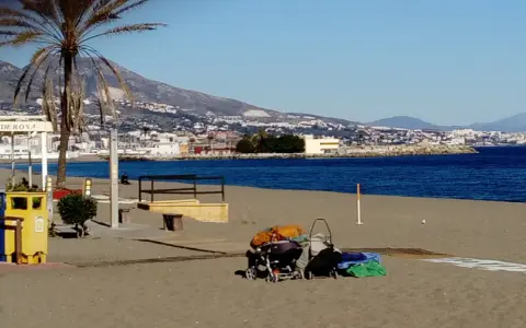 Sonniger fuengirola strand mit Palmen, Bergen und dem Meer im Hintergrund. Kinderwagen und Gepäck liegen am Sand.