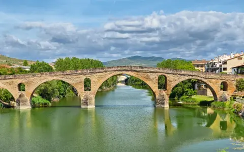 Der historische Puente la Reina überspannt mit seinen steinernen Bögen einen grünen Fluss. Dahinter erstreckt sich eine malerische Stadtkulisse.