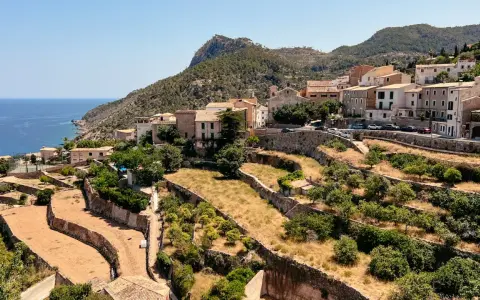 Terrassierte Felder und Häuser schmiegen sich an die Hänge des Tramuntana Gebirges, mit dem blauen Meer im Hintergrund.