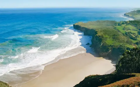 Atemberaubende Küstenlandschaft am Camino del Norte. Grüne Hügel treffen auf das azurblaue Meer mit sanften Wellen, die auf einen einsamen Sandstrand treffen.