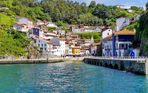 Bunte Häuser schmiegen sich an einen grünen Hang in Cudillero, Spanien. Das türkisfarbene Wasser des Hafens spiegelt die malerische Kulisse wider.