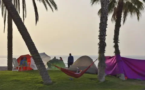 Entspannter Camping-Urlaub an der Costa del Sol. Palmen, Zelte, Sonnenschirme und eine Hängematte am Strand.