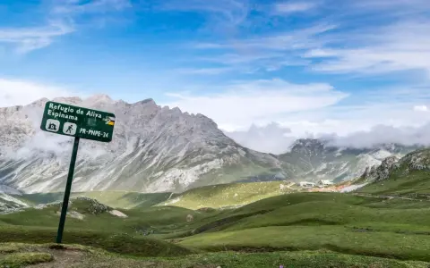 Wanderwegschild weist den Weg zum Refugio de Aliva und Espinama in den **Lagos de Covadonga**. Grüne Hügel und zerklüftete Berge unter blauem Himmel.