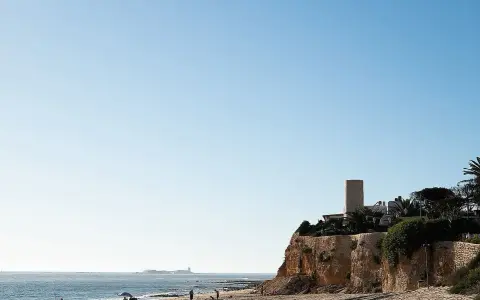 Sonniger Tag am **Playa La Barrosa**. Menschen genießen den Strand, während im Hintergrund eine Festung am Meer thront.