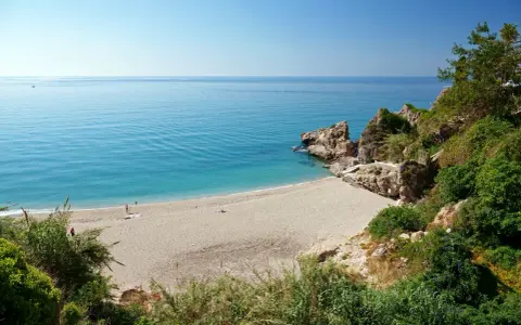 Playa de Burriana: Ein sonniger Strand mit türkisfarbenem Wasser, Felsen und üppiger Vegetation. Einige Menschen genießen die Ruhe.