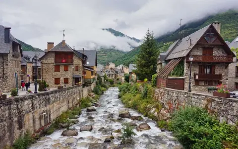 Ein rauschender Fluss schlängelt sich durch das malerische Val d'Aran, gesäumt von traditionellen Steinhäusern und grünen Bergen.