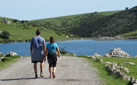 Ein Paar wandert auf einem Schotterweg am Ufer eines Sees. Grüne Hügel und Felsen prägen die **Spanien Natur**.