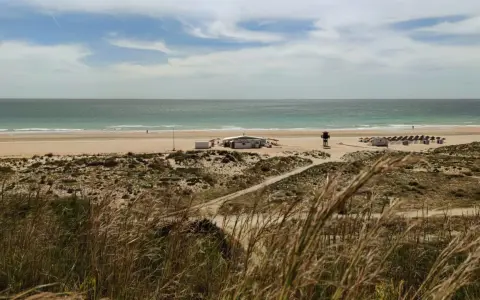 Strand von Conil de la Frontera mit Dünen, einem Strandcafé und Sonnenschirmen. Das Meer ist ruhig, der Himmel ist bewölkt.