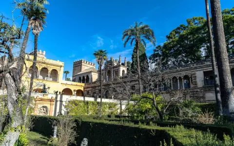 Sonniger Tag im Alcázar von Sevilla. Palmen ragen über kunstvoll verzierte Gebäude und gepflegte Gärten.