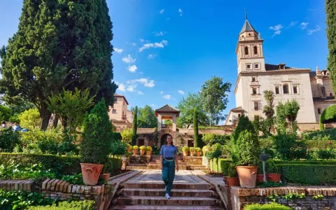 Frau spaziert durch einen üppigen Garten mit Zypressen und einem Turm im Hintergrund. Ein schöner Ort für einen Tagesausflug, vielleicht sogar von Malaga aus.