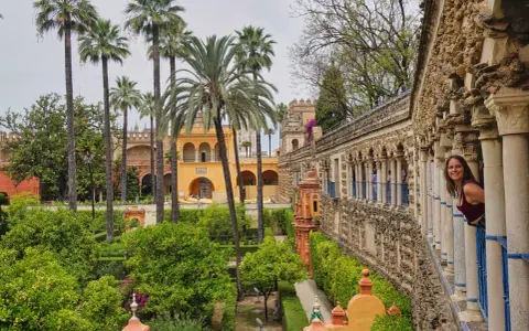 Frau blickt lächelnd aus einem Säulengang auf üppige Gärten mit Palmen. Ein versteckter Blick auf Sevilla, der Lust auf mehr macht. #sevilla #geheimtipps