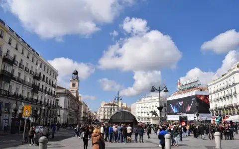 Menschenmassen beleben den Platz Puerta del Sol in Madrid unter einem blauen Himmel mit Wolken. Der Uhrturm und das Tio Pepe Schild sind sichtbar.
