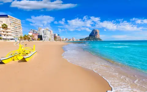 Sonne, Strand und Meer in Calpe, Spanien. Ein gelbes Wasserfahrrad wartet am Sandstrand, im Hintergrund die Stadt und der markante Felsen Peñón de Ifach. Ein echter Geheimtipp!