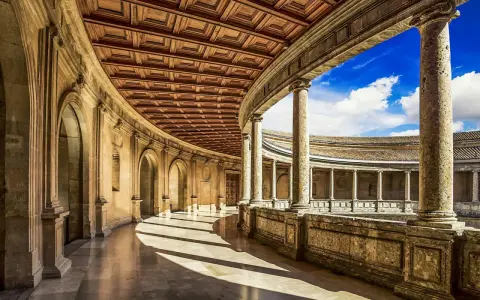 Ein sonnenbeschienener Arkadengang mit kunstvoller Holzdecke und Säulen, der zum Innenhof der Alhambra Moschee führt.