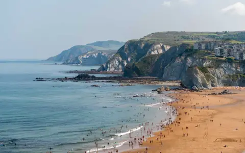 Ein belebter Strand mit vielen Menschen, die im Meer schwimmen und surfen, vor einer dramatischen Felsküste. Das Bilbao Meer lädt zum Entspannen ein.