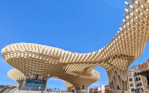 Der Metropol Parasol in Sevilla ragt mit seiner Holzkonstruktion in den blauen Himmel. Menschen steigen die Treppen hinauf.