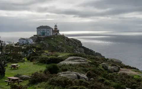 Leuchtturm und Gebäude auf felsiger Küste bei Finisterre, Spanien. Grauer Himmel über dem Meer.