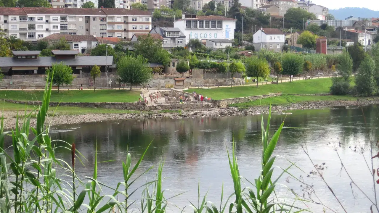 Menschen entspannen sich an einem Flussufer in Ourense, Spanien. Im Hintergrund sind Geb&auml;ude und gr&uuml;ne H&uuml;gel zu sehen.