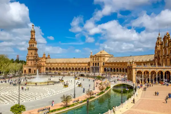 Der beeindruckende Sevilla Palast mit seinen Türmen und Arkaden, umgeben von einem Kanal und belebtem Platz mit Brunnen und Kutschen.