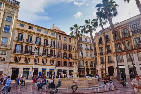 Sonniger Platz in der **Malaga Altstadt** mit einem Brunnen, Palmen und bunten Gebäuden. Menschen flanieren und genießen die Atmosphäre.