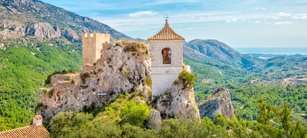 Das Castell de Guadalest thront auf einem Felsen, umgeben von grünen Bergen und dem blauen Meer in der Ferne. Ein Glockenturm ragt empor.