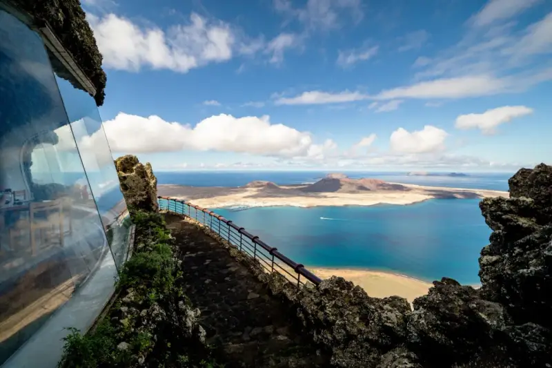 Blick vom Mirador del Río auf das Tal der tausend Palmen Lanzarote. Blaue See, Inseln und eine Hütte mit Glasfront.