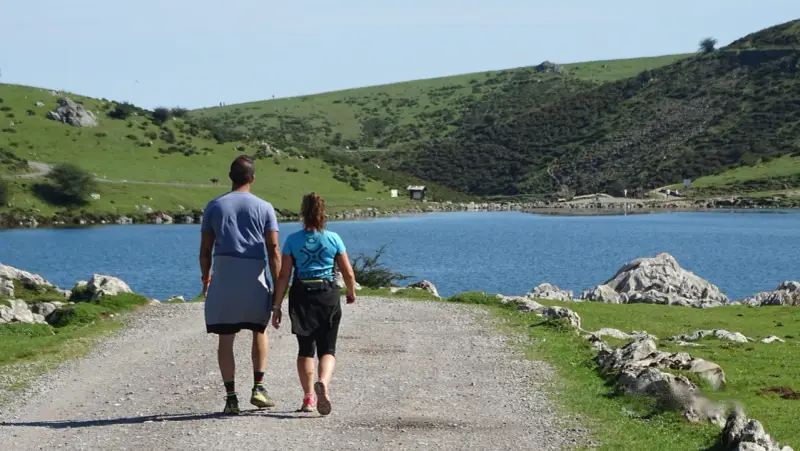 Ein Paar wandert auf einem Schotterweg am Ufer eines Sees. Grüne Hügel und Felsen prägen die **Spanien Natur**.